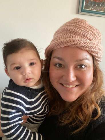 L'image montre une femme portant un chapeau rose et tenant un bébé devant un mur blanc orné d'un tableau. La femme a de longs cheveux bruns et le bébé porte un pull à rayures.