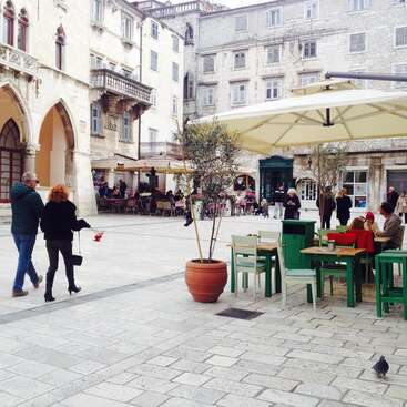 L'image représente un charmant espace extérieur sur une place historique, avec des tables, des chaises et des parasols, entouré de bâtiments ornés et de gens qui se promènent.