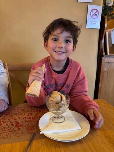Un enfant souriant, vêtu d'un pull rose, est assis à une table, heureux de tenir un cornet de glace et un bol de glace en verre devant lui.