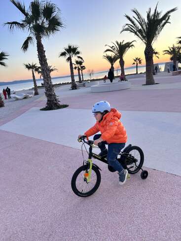 Un jeune enfant vêtu d'une veste orange roule sur un vélo à roulettes le long d'une promenade bordée de palmiers au coucher du soleil, près de la mer, en profitant du plein air.