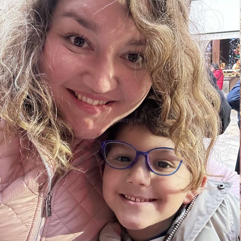 Une femme et un enfant sourient chaleureusement à la caméra. La femme a les cheveux bouclés et l'enfant porte des lunettes bleues. Tous deux sont vêtus de vestes légères.