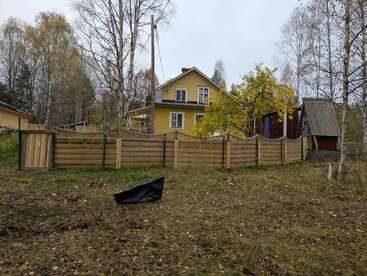 Ein gelbes Haus mit weißer Zierleiste steht hinter einem Holzzaun. Herbstliche Bäume umgeben den Hof, und eine schwarze Plane liegt auf dem grasbewachsenen Boden.