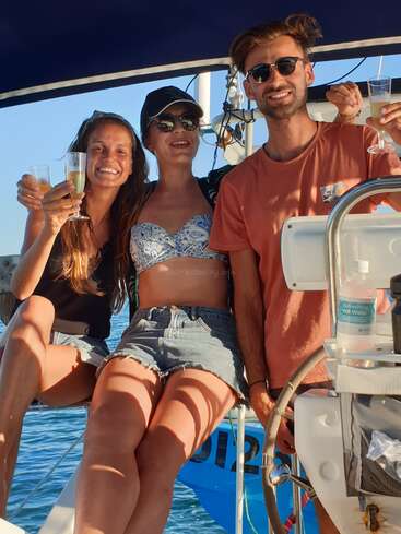 The image depicts three individuals on a boat, each holding a glass of champagne, with the man wearing an orange shirt and the woman in the middle wearing a bikini top.