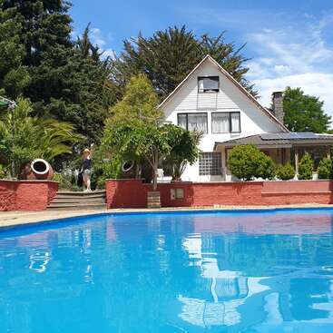 Das Bild zeigt ein weißes Haus mit einem großen Pool davor, umgeben von Bäumen und Büschen, an einem sonnigen Tag mit blauem Himmel.