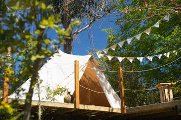 A white canvas tent sits on a wooden deck surrounded by trees, with festive triangular bunting overhead. Sunlight filters through, creating a peaceful, rustic outdoor scene.
