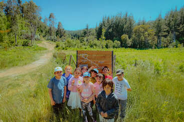 A group of children stands together in a grassy field on a sunny day, with a wooden map sign behind them and trees in the background.