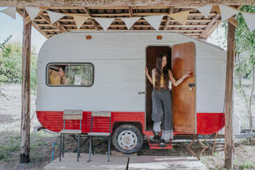 Two young women are at a vintage red and white camper under a wooden shelter. One stands in the doorway, the other looks out the window. Festive atmosphere.