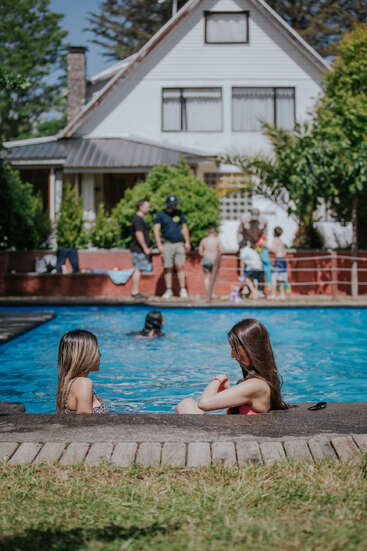 Two women relax at the edge of a pool, children and adults play in the background, and a large house sits behind lush green trees.