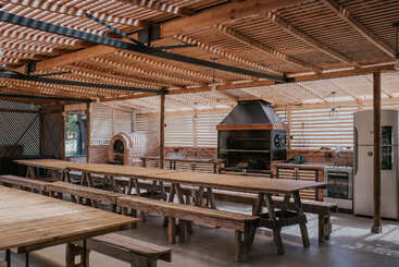 This image shows a rustic, open-air kitchen and dining area with wooden tables, benches, a large grill, pizza oven, refrigerator, and abundant natural light. Inviting space.