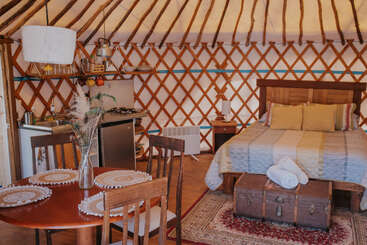This cozy yurt interior features a neatly made bed, wooden furniture, small kitchenette, vintage trunk, and a dining area, creating a warm, rustic atmosphere.