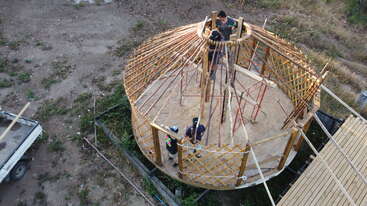 This image shows four people constructing a circular wooden structure with an open roof. The structure appears to be a yurt, surrounded by dirt and sparse vegetation.