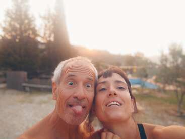 A imagem mostra um homem e uma mulher posando para uma selfie, com o homem colocando a língua para fora e a mulher sorrindo. Eles estão do lado de fora, em um ambiente natural.