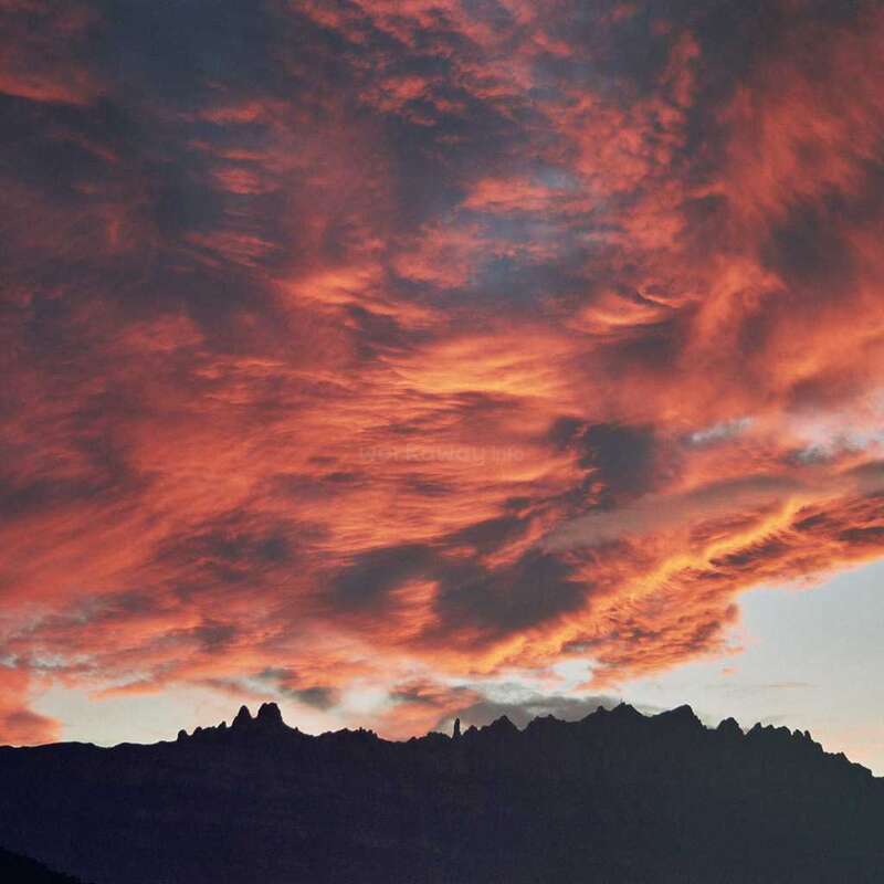 A imagem retrata um pôr do sol de tirar o fôlego sobre uma cadeia de montanhas, com nuvens laranja e rosa vibrantes iluminando o céu acima da silhueta escura das montanhas.