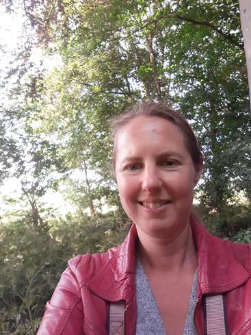 The image depicts a woman with brown hair, wearing a red leather jacket and grey shirt, standing in front of trees and bushes on a sunny day.