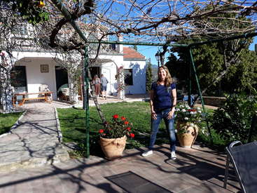 The image depicts a woman standing on a patio in front of a house, surrounded by potted plants and trees, with a serene and peaceful atmosphere.