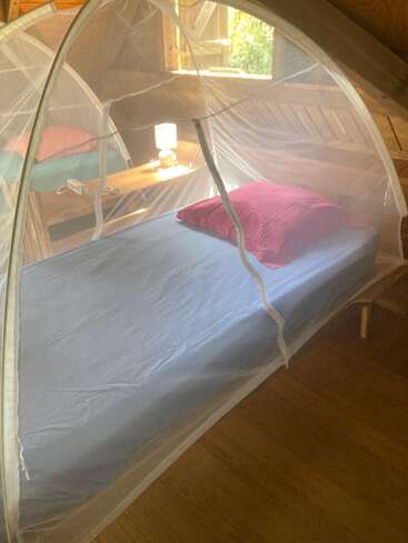 Ein gemütliches Bett mit blauem Laken und rotem Kissen ist von einem Netzzelt umgeben. Daneben steht ein hölzerner Nachttisch mit einer Lampe und einem Buch.