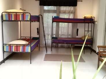 The image depicts a room with two bunk beds, each featuring a ladder and striped bedding, accompanied by a rug and a chair, set against a white wall and floor.