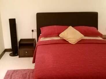 The image depicts a bedroom with a bed, nightstand, and speaker, featuring a red comforter, pink pillows, and a pink rug on a white floor against a white wall.