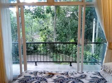 The image depicts a serene bedroom with a large window overlooking a lush forest, featuring a bed with a blue and white comforter and sheer curtains on either side.
