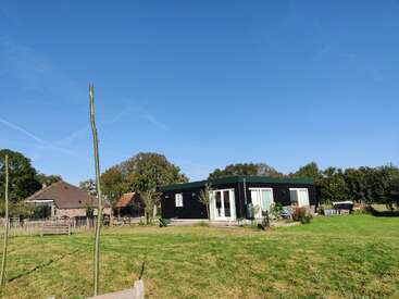 Une maison moderne noire de plain-pied se dresse dans une cour spacieuse et herbeuse, sous un ciel bleu clair. Elle est entourée d'arbres, d'une clôture en bois et de maisons voisines.