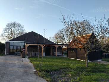 Dans un cadre rural paisible, une grande maison en briques avec un toit en tuiles, une petite cabane en bois, des arbres dénudés, de l'herbe verte et un ciel dégagé.