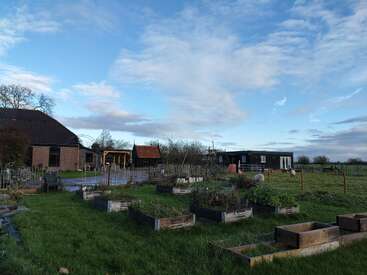 Scène rurale avec des potagers surélevés, un champ herbeux, des moutons en train de paître, plusieurs bâtiments agricoles, des arbres et un ciel bleu clair rempli de nuages épars. Atmosphère paisible.
