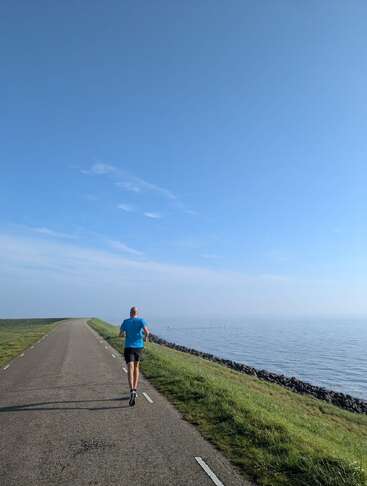 Un homme en chemise bleue fait son jogging seul sur une route pavée au bord de l'eau, sous un vaste ciel bleu clair avec de l'herbe verte autour.