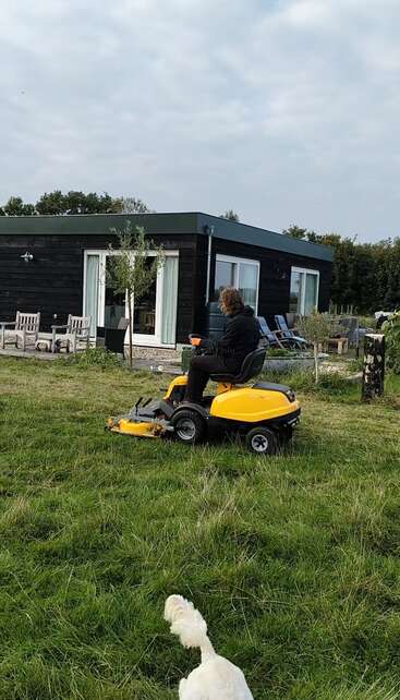 Une personne utilise une tondeuse à gazon jaune pour couper l'herbe devant une maison moderne noire. Un chien blanc marche sur l'herbe à proximité, sous un ciel nuageux.
