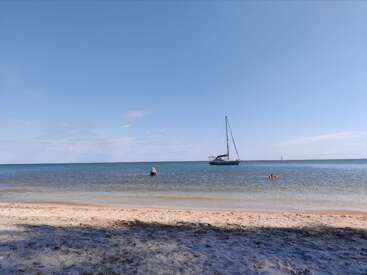 Uma cena de praia tranquila com duas pessoas nadando no mar, um veleiro ancorado nas proximidades, ondas suaves, costa arenosa e um céu azul claro.