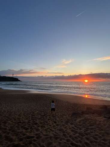 Ein einsames Kind steht bei Sonnenuntergang an einem Sandstrand und schaut aufs Meer. Auf der Klippe in der Ferne steht ein Leuchtturm, der Himmel ist in Farben getaucht.
