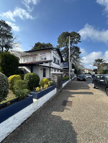 Ein cooles Hotel namens „Chez Benat” liegt in einer sonnigen Straße mit ordentlich geschnittenen Büschen, geparkten Autos und hohen Bäumen unter blauem Himmel.