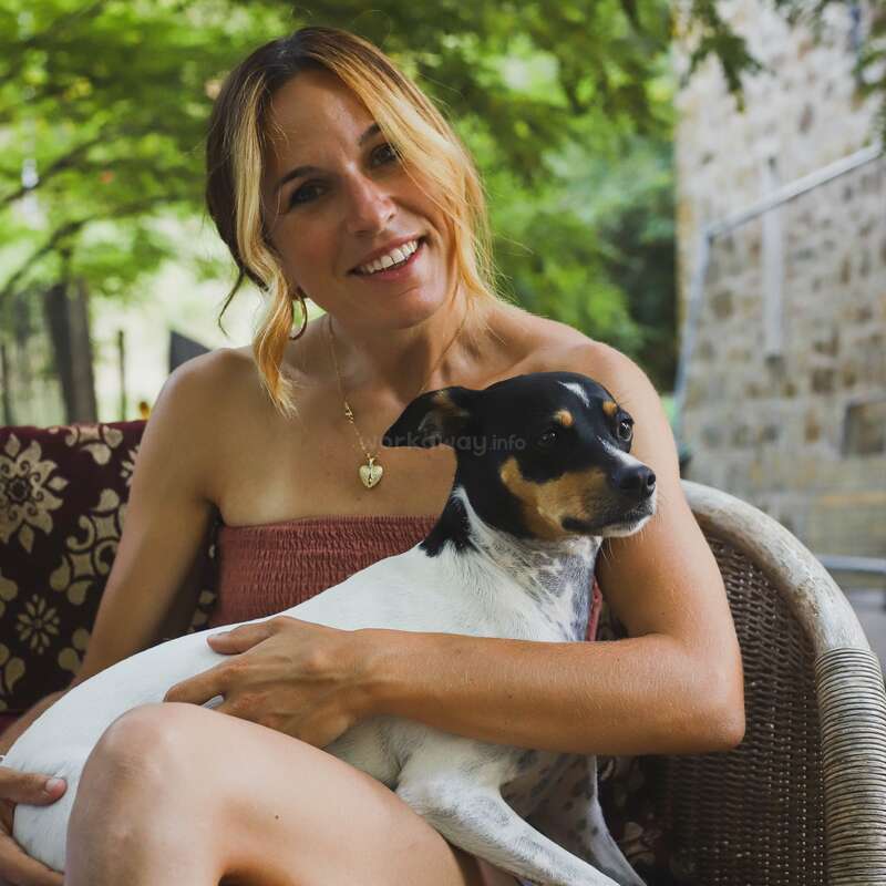 Eine lächelnde Frau mit blondem Haar sitzt im Freien auf einem Korbstuhl und hält liebevoll einen kleinen schwarz-weiß-braunen Hund auf ihrem Schoß. Üppiges Grün umgibt sie.