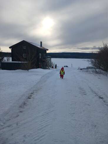 Un niño con ropa de invierno de colores vivos camina por un sendero cubierto de nieve cerca de una casa de madera. El sol brilla a través de un cielo nublado, creando una escena serena.