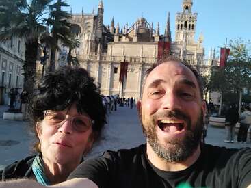 Ein fröhlicher Mann und eine Frau machen ein Selfie vor einer historischen, kunstvollen Kathedrale unter strahlend blauem Himmel, umgeben von Palmen und Touristen.