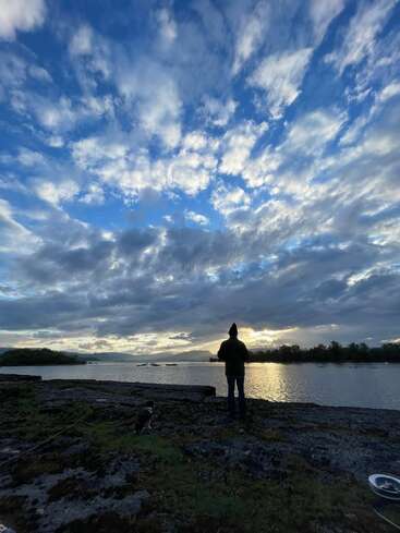 A person stands by a serene lakeshore at sunset, with dramatic clouds overhead. A dog sits nearby, both gazing across the reflective, tranquil water. Peaceful moment.