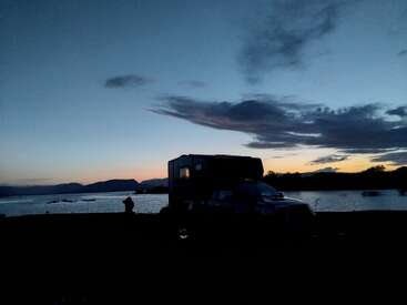 A truck and camper are silhouetted against a colorful sunset by a serene lake, with dramatic clouds and mountains in the distance, evoking peaceful solitude.