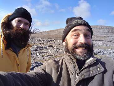 Two bearded men in jackets and hats smile for a selfie outdoors on a rocky, barren landscape under a blue sky with scattered clouds.