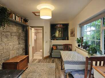 This cozy, well-lit living space features a stone wall, a small sofa, dining table, bookshelves, plants, wooden furniture, and a modern bathroom visible.