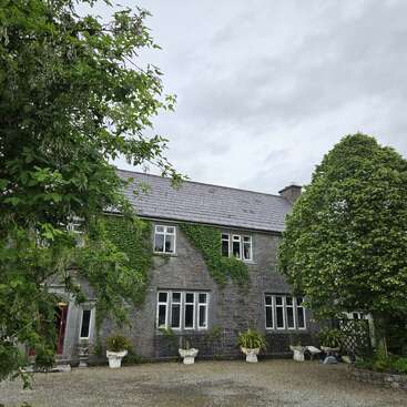 The image shows a charming stone house with ivy-covered walls, surrounded by lush green trees and potted plants, under an overcast sky. Peaceful and inviting atmosphere.