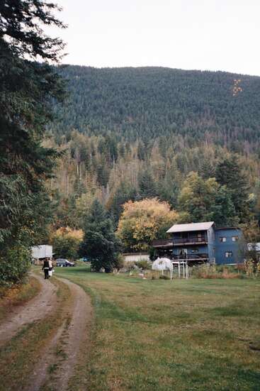 Uma casa azul fica entre árvores altas e vegetação, perto de uma montanha. Um caminho de terra sinuoso leva a duas pessoas que caminham tranquilamente pelo quintal.