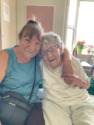 Duas mulheres, uma mais velha e outra mais jovem, estão sentadas bem juntas, sorrindo calorosamente. A mulher mais velha usa óculos. A luz natural e as flores iluminam o ambiente aconchegante.