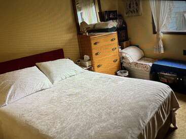 Um quarto aconchegante com uma cama bem arrumada, uma cômoda de madeira, um pequeno sofá-cama, uma janela com cortinas de renda e vários objetos pessoais, criando um ambiente acolhedor e convidativo.