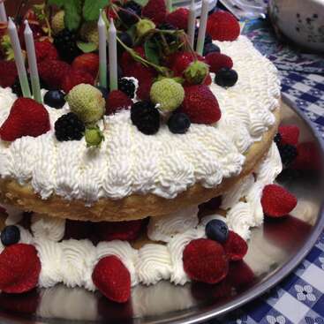 A two-layer cake topped with whipped cream, fresh strawberries, blueberries, blackberries, and green berries. White candles are inserted on top. Cake is on a silver platter.