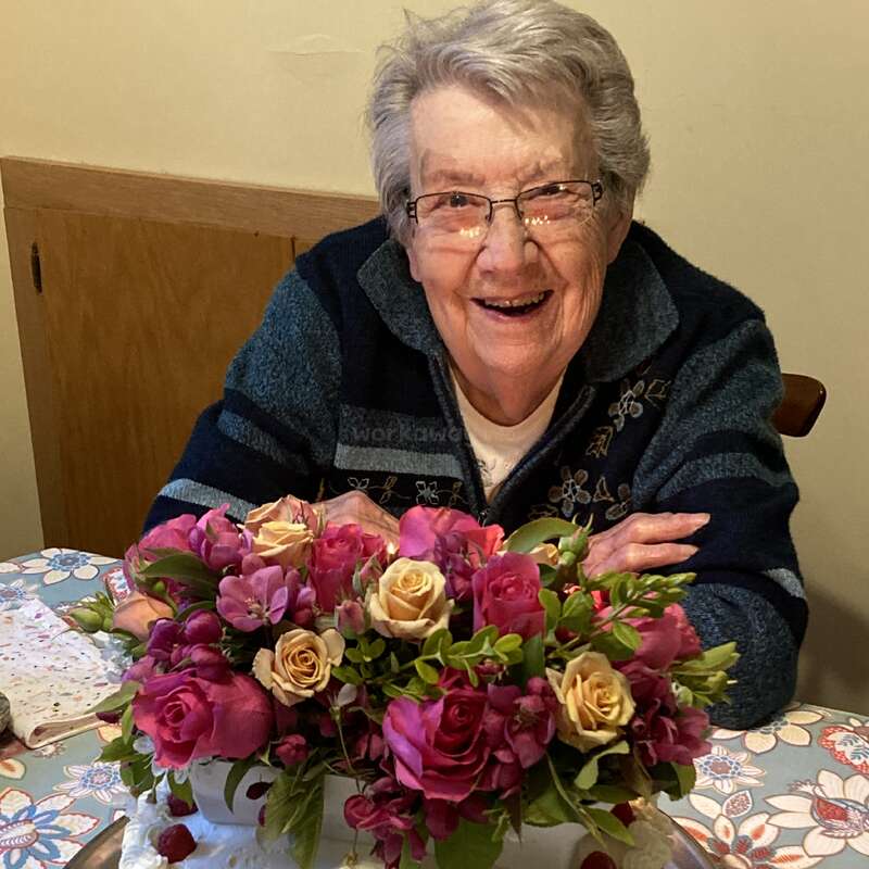 Uma senhora idosa, de cabelo curto e grisalho e usando óculos, sorri calorosamente, sentada a uma mesa decorada com um buquê colorido de rosas e outras flores.