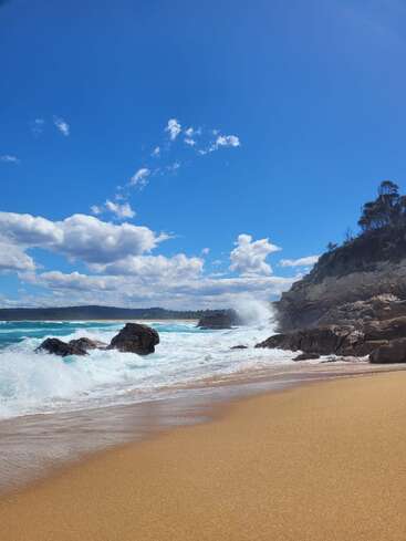Golden sand stretches along the beach, waves crash against rocky cliffs, turquoise water sparkles under a vibrant blue sky filled with fluffy white clouds. Nature's beauty.