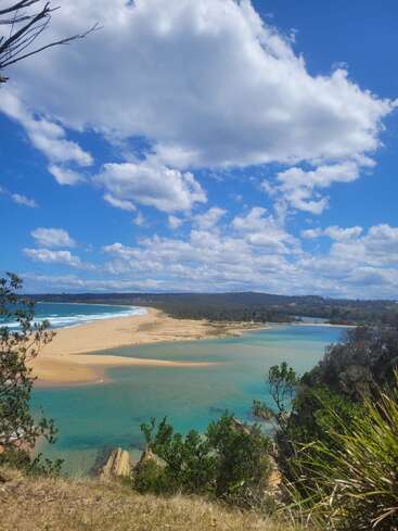 This image shows a beautiful coastal scene with turquoise water, sandy beaches, lush greenery, and a blue sky filled with fluffy white clouds on a sunny day.