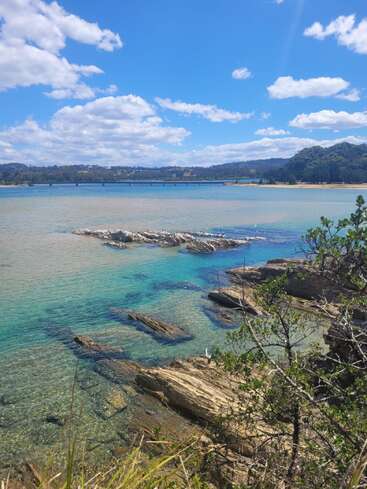 A breathtaking coastal scene with turquoise water, rocky shoreline, clear blue sky, scattered clouds, distant forested hills, a long bridge, and lush greenery in the foreground.