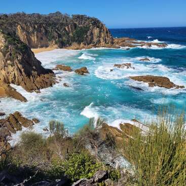 Clear turquoise waves crash against rugged rocks and golden cliffs under a cloudless blue sky, surrounded by lush greenery. A secluded, pristine coastal paradise.