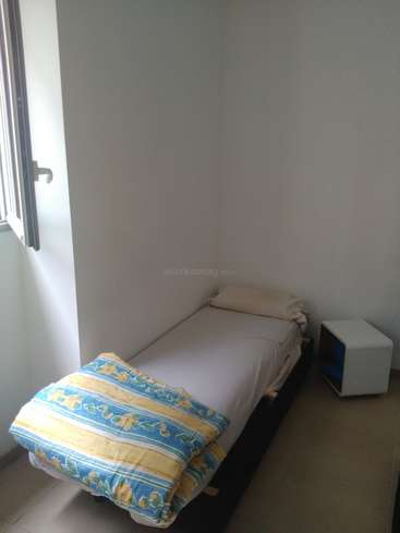 The image depicts a small, minimalist room with a single bed, white walls, and a window, featuring a blue and yellow blanket and a small white nightstand.