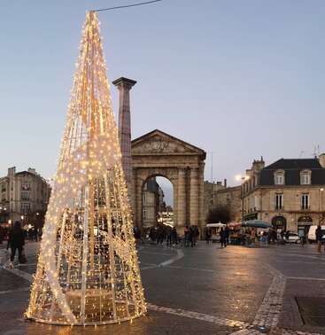 The image depicts a festive scene with a tall, illuminated Christmas tree in the foreground, set against a backdrop of a stone archway and historic buildings.
